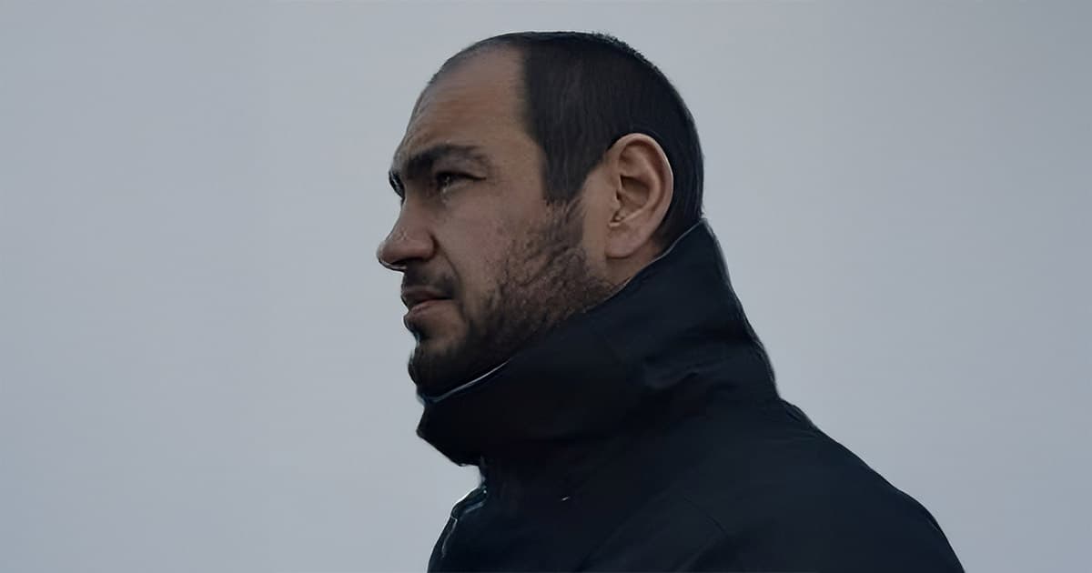 A man with short dark hair and a beard, wearing a black jacket, looks to the left against a plain, light gray background.