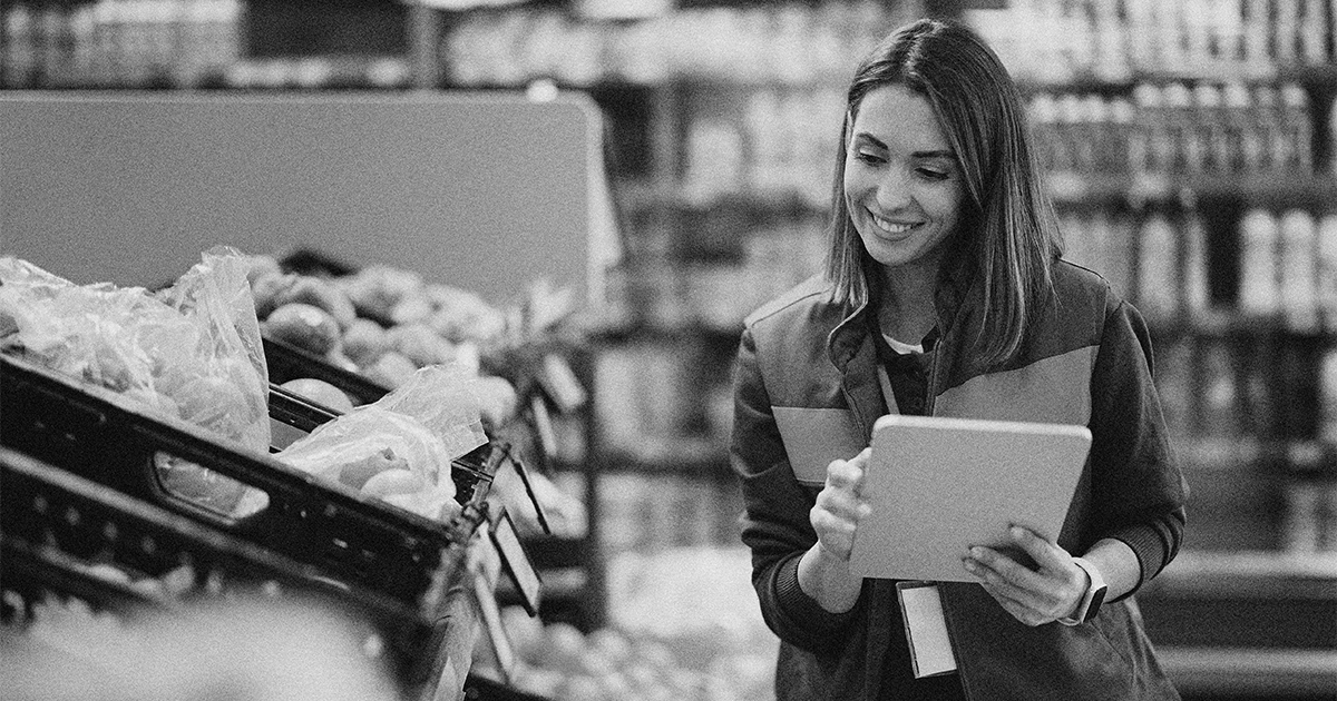 A woman wearing a uniform smiles while holding a tablet and checking produce in a grocery store aisle.