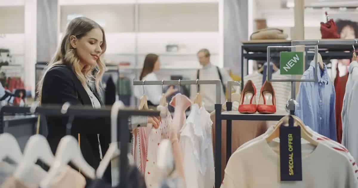 A woman shops for clothes in a store, browsing through garments on a rack. A Special Offer tag and a New sign are visible, with red high heels displayed nearby. Other shoppers are seen in the background.