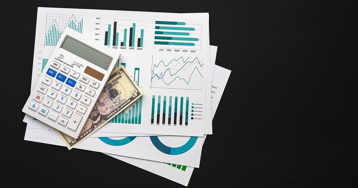 A calculator and dollar bills resting on printed financial charts with various bar and line graphs, all set against a black background.