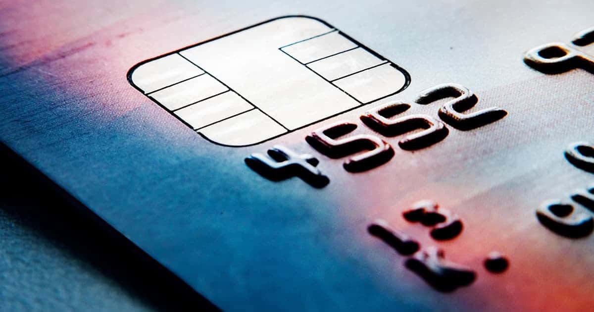 Close-up of a credit card showing a metallic chip and raised numbers on a blue and red gradient background. The card details are partially visible and out of focus.