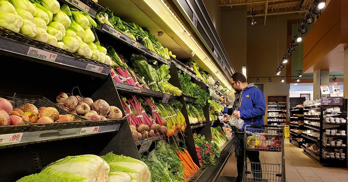 A man wearing glasses and a blue jacket shops for fresh vegetables in a grocery store produce aisle, placing items into his shopping cart filled with groceries.