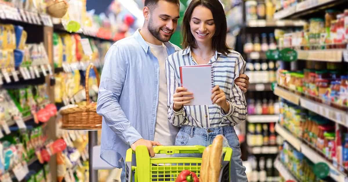 A smiling couple stands in a grocery store aisle, looking at a shopping list together while pushing a cart filled with groceries, including a baguette and fresh produce.