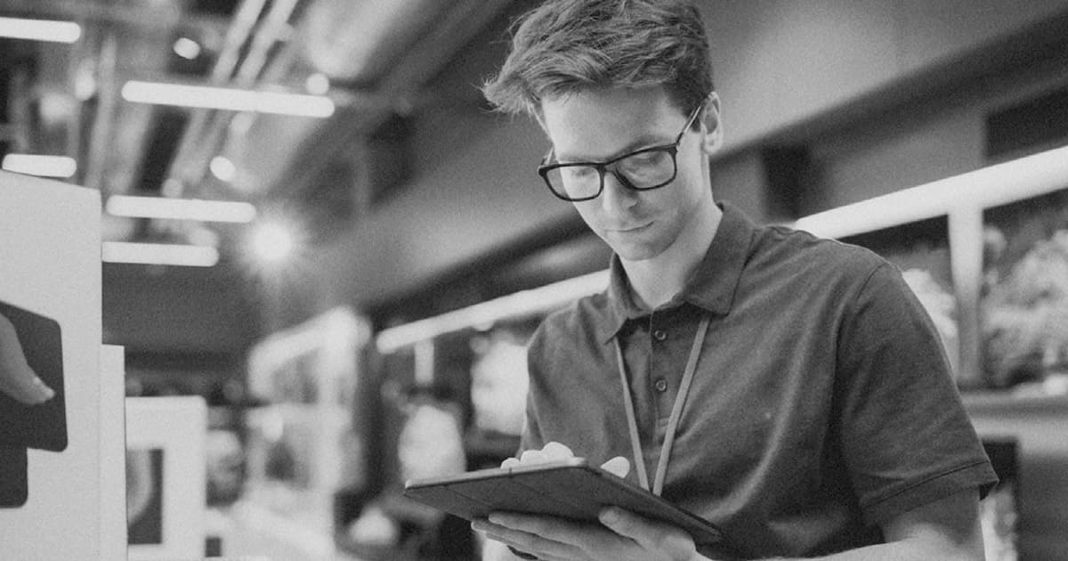 A young man wearing glasses and a polo shirt is standing in a store, using a tablet. He appears to be focused on his work.