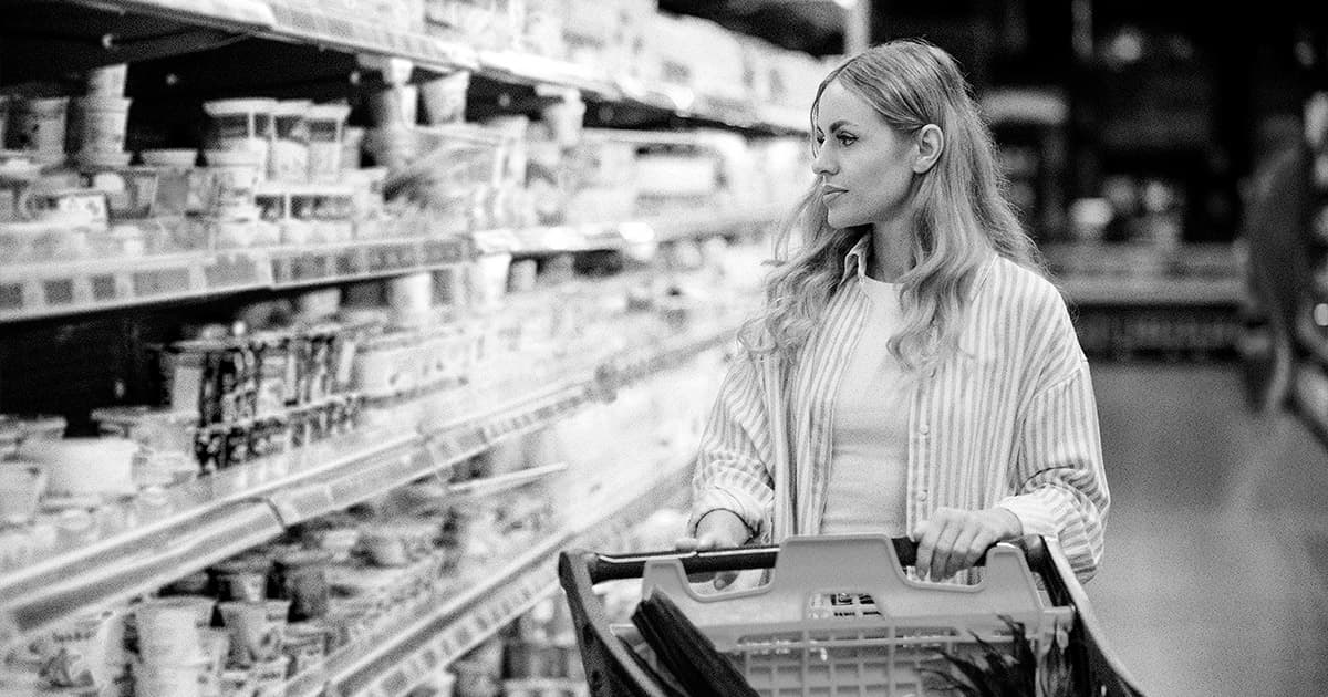 A woman with long hair is pushing a shopping cart and looking at products on a grocery store shelf. The image is in black and white.