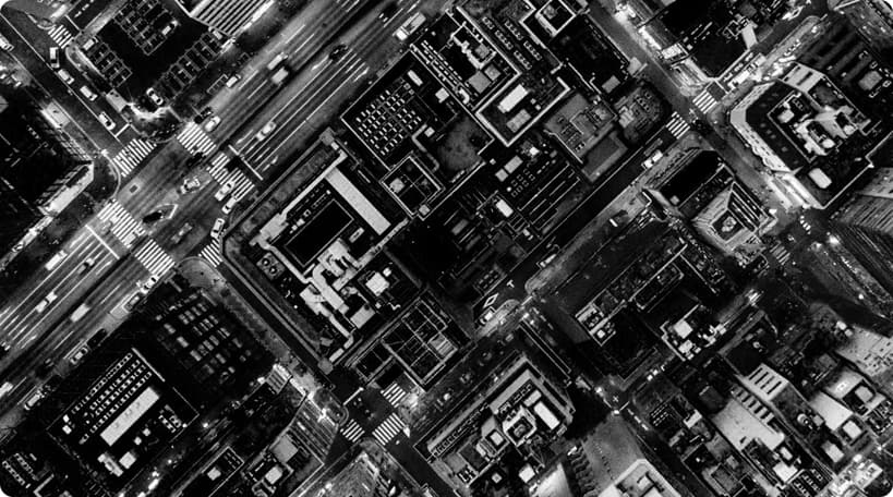 A black and white aerial photo of a city intersection, showing several streets crossing at right angles surrounded by densely packed buildings and illuminated crosswalks.