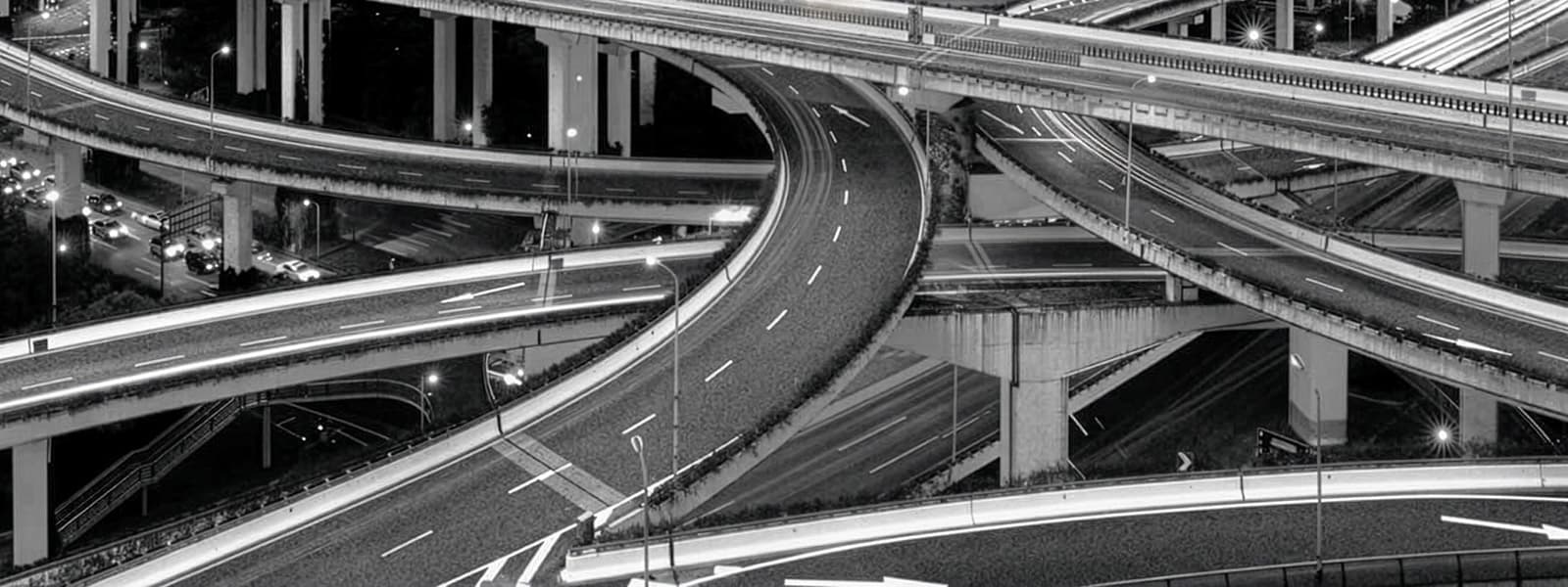 A black and white photo of a complex multi-level highway interchange, featuring several curving overpasses, elevated roads, and visible traffic lanes with a few cars in the background.