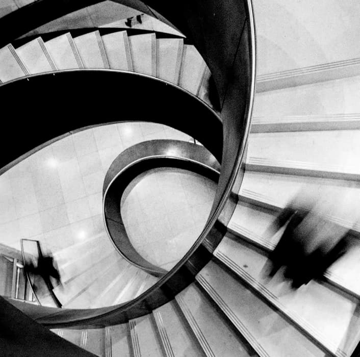 Black and white photo of a spiral staircase seen from above, with blurred figures moving up and down the steps, creating a dynamic and abstract visual effect.