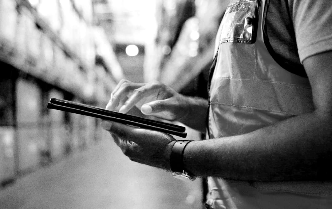 A person wearing a safety vest uses a tablet in a warehouse aisle, with shelves full of boxes blurred in the background.