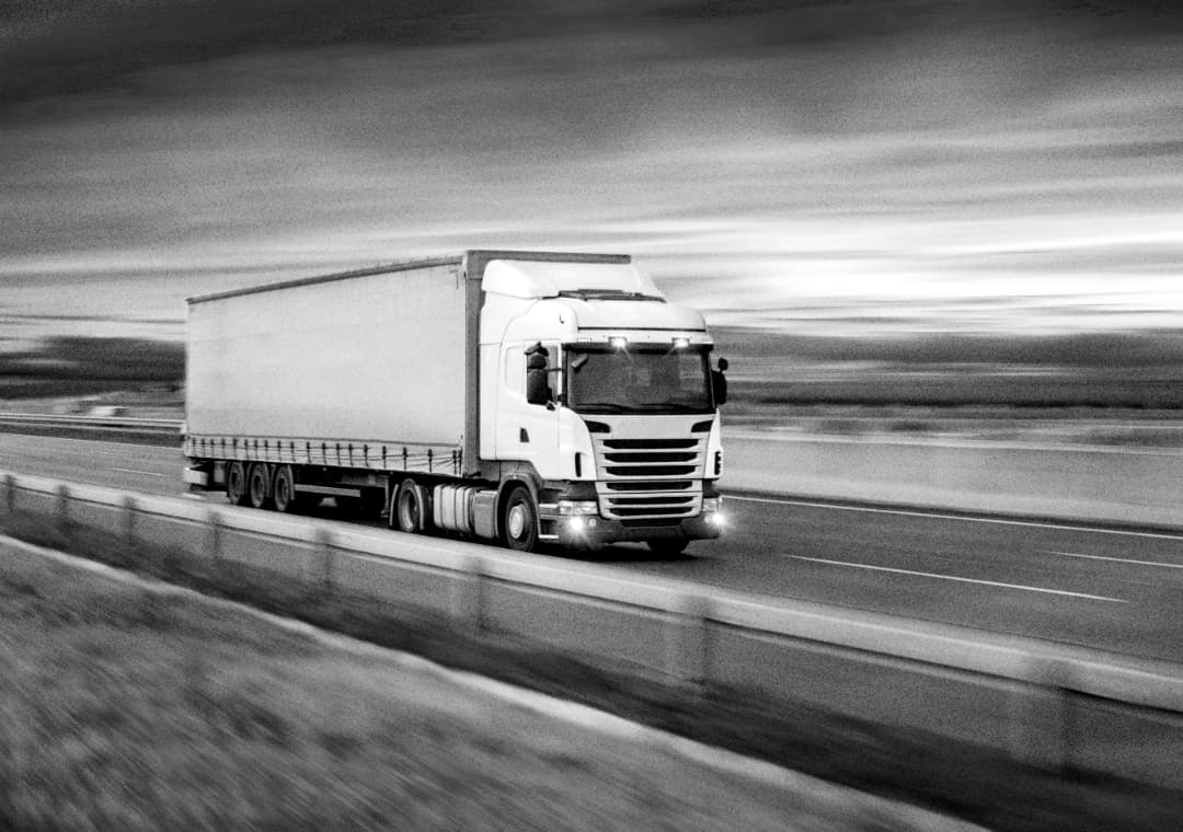 A large white semi-truck drives quickly along a highway, with motion blur in the background indicating speed; the scene is in black and white.