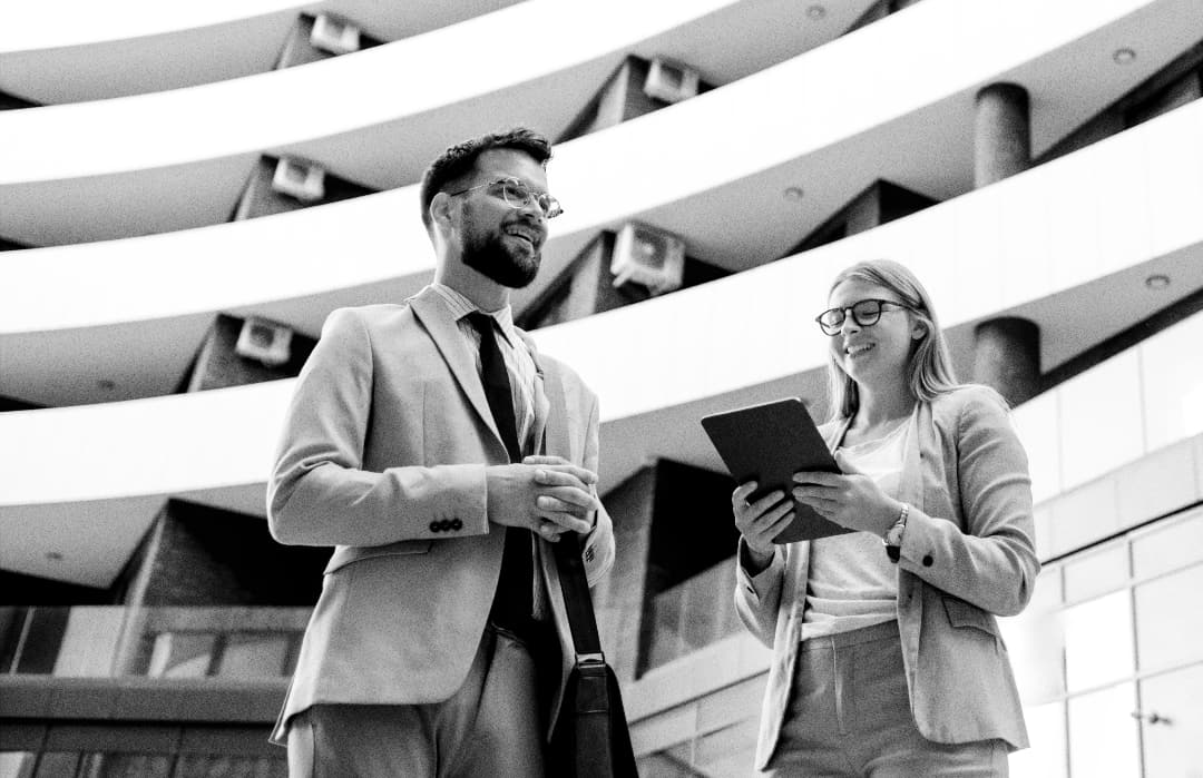 Two business professionals in suits stand outside a modern, curved office building. The woman holds a tablet and smiles, while the man, wearing glasses, looks on, both appearing engaged in conversation.