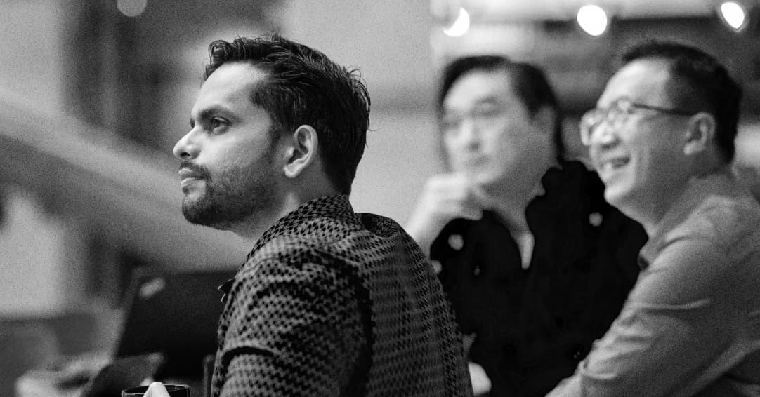 A man wearing glasses and a light-colored shirt sits on a couch, gesturing with his right hand while smiling in a modern, well-lit room. The image is in black and white.