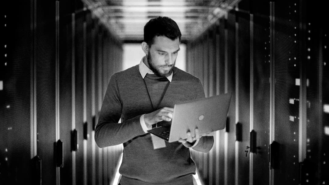A man stands in a server room corridor, looking at and typing on a laptop. Rows of server racks and dim lighting emphasize a high-tech, data-centric environment.