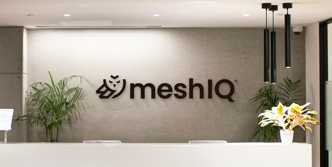 A modern reception area with the meshIQ logo on the wall, two green plants on either side of a white desk, and three black pendant lights hanging from the ceiling.