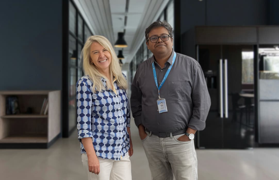 Two people stand smiling in a classroom with rows of desks, each desk holding a white gift bag with green tissue paper and an owl logo. The room has white walls and black chairs.