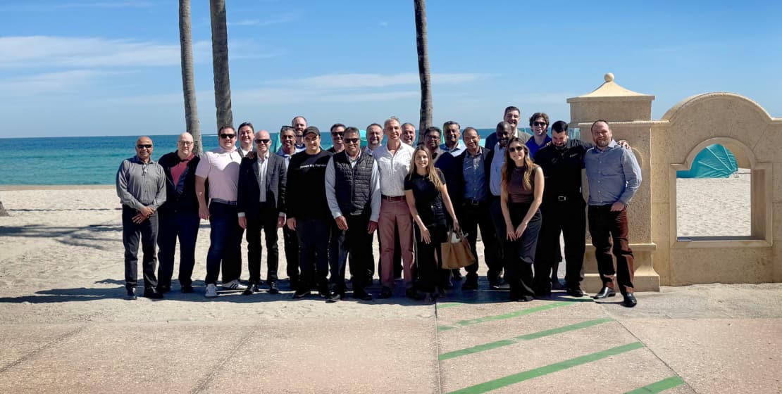A group of people pose together on a sunny beach boardwalk, with the ocean, blue sky, and palm trees in the background. Some people wear business attire, while others are dressed casually.