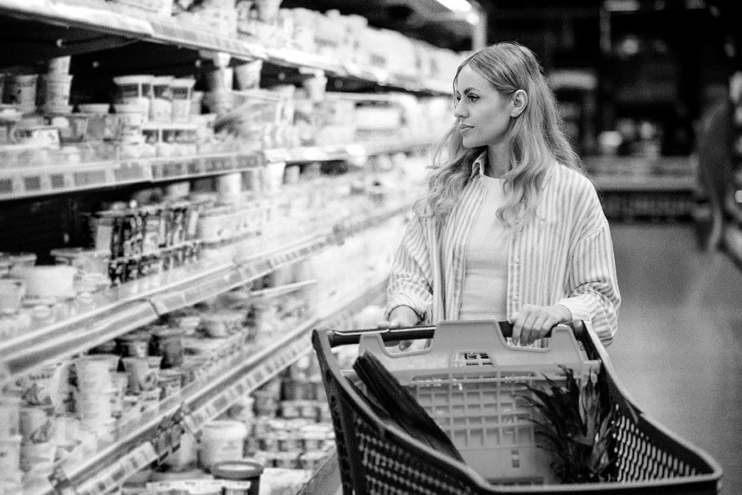 A woman with long hair pushes a shopping cart and looks at dairy products on shelves in a grocery store. The image is in black and white.