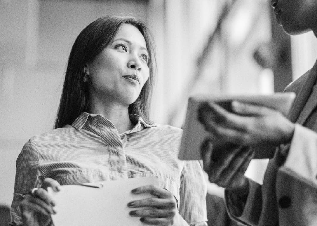 A woman listens attentively to a person holding a tablet or notebook. The image is black and white, and both individuals appear to be engaged in conversation.